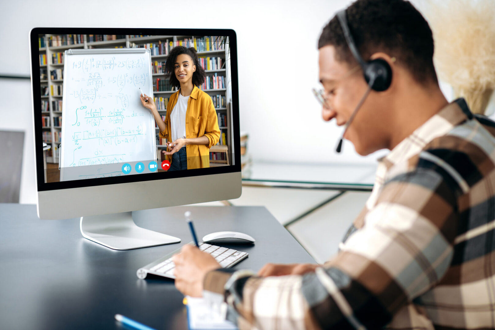 Distant learning, webinar, online lecture. Hispanic clever male student, sits at work desk, uses computer, listening to an online lecture, taking notes, female african american teacher on the screen Online tutoring session with whiteboard explanation.